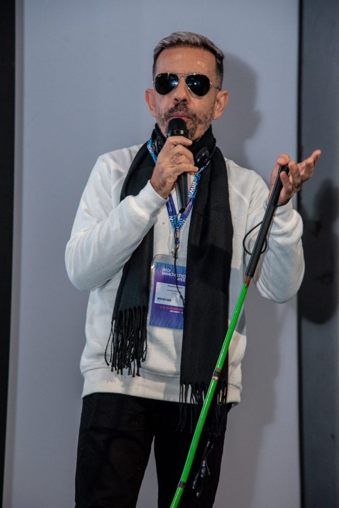 Foto vertical de  Felipe Monteiro na Rio Inovation Week 2024.
Felipe é branco, cabelos pretos, curtos, corte no estilo militar, com as laterais mais curtas que o topo, onde as pontas estão descoloridas. Rosto alongado, testa alta, barba por fazer e bigode também curtos e pretos. 
Usa suéter branco, calça preta e óculos de sol triangular dourado, cachecol azul marinho e um crachá azul pendurado no pescoço. 
Os braços estão flexionados. O direito para cima, na diagonal a frente do dorso. A mão direita, na altura do pescoço, segura um microfone a frente da boca. 
O braço esquerdo em linha diagonal ascendente, ao lado e a frente do corpo, com a mão na altura do ombro, com os dedos estendidos, meio abertos. O polegar flexionado segura uma bengala verde, apoiada na palma da mão descendo em diagonal na frente das pernas e em direção ao piso. As pernas estão alongadas, levemente abertas e ocultas a partir dos joelhos.  
Atrás dele, a direita, parte de sua sombra projetada na parede de fundo cinza.
Roteiro: Nadia Moreno
Consultoria: Felipe Monteiro
