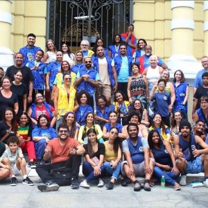 Foto vertical de Felipe Monteiro com grupo de Ações educativas da ciência móvel no Instituto Nacional de Educação de Surdos (INES) em março de 2025. Ele é branco, cabelos pretos, curtos, corte no estilo militar, com as laterais mais curtas que o topo, onde as pontas estão descoloridas. Rosto alongado, testa alta, barba e bigode cheios, também curtos e pretos. Usa um colete azul royal, com botões brancos sobre uma camiseta azul celeste e calça preta, óculos de sol triangular dourado. Os braços estão flexionados paralelamente ao dorso. A mão direita, apoiada na esquerda, que segura uma bengala na frente das pernas alongadas. Ao Redor dele, muitas pessoas, sentadas, abaixadas e em pé, em pose para foto. São homens e mulheres, brancos e negros. Eles estão em uma escadaria a frente ao prédio do Instituto Nacional de Educação de surdos – INES. Um edifício histórico amarelo com um portão em ferro preto gradeado e com arabescos ao centro. Nas laterais, à frente do portão, nas duas extremidades colunas também amarelas com detalhes em branco. Parte do grupo está ocupando o espaço à frente das colunas e no piso à frente do final da escada. Algumas pessoas usam colete similar ao de Felipe, outras usam um colete amarelo e dentre as sem colete há um grupo de camiseta preta. Roteiro: Nadia Moreno Consultoria: Felipe Monteiro 