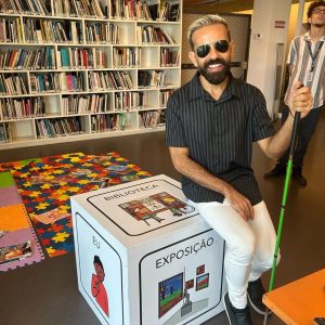 Foto vertical de Felipe Monteiro conhecendo a comunicação alternativa da biblioteca do Museu de Arte do Rio (MAR), em março de 2025. Ele é branco, cabelos pretos, curtos, corte no estilo militar, com as laterais mais curtas que o topo, onde as pontas estão descoloridas. Rosto alongado, testa alta, barba e bigode cheios, também curtos e pretos. Esboça um sorriso, mostrando os dentes brancos e alinhados. Usa camisa de manga curta, cinza chumbo, com listras verticais pretas e botões pretos, calça branca, tênis preto com solado branco, óculos de sol triangular dourado e uma aliança no dedo anelar da mão esquerda. Os braços e pernas estão flexionados. O direito em diagonal para baixo, apoiado na perna direita, a mão, do mesmo lado, alongada, repousa entre as duas pernas. O esquerdo flexionado para cima. A mão segura uma bengala verde. A perna direita, um pouco mais elevada que a esquerda. Os pés flexionados, com as pontas no piso e os calcanhares em suspenso. Ele está sentado, apoiado na quina esquerda de um cubo branco. O Bloco contém imagens de comunicação alternativa aumentativa aplicadas em suas faces. Na lateral direita, o símbolo para eu (o pictograma de um menino com o dedo apontado para si mesmo). Na face frontal, outro pictograma que representa exposição (a representação de uma sala, no encontro de duas paredes, com quadros e uma estátua no piso. Na face superior, o pictograma para biblioteca, (duas pessoas, uma delas em cadeira de rodas, sentadas frente a frente, em uma mesa. Ao fundo uma estante com livros). Atrás do cubo, sobre o piso preto, parte de dois tapetes de EVA, com revistas espalhadas. Um deles formado por quadrados de encaixe, nas cores lilás, laranja e verde. O outro, por peças no formato de quebra-cabeças, em vermelho, azul, laranja e amarelo. Uma fileira com 4 peças retangulares de encaixe, nas cores laranja, verde, vermelho e amarelo, com letras em preto, estão ao final do tapete. A esquerda e um pouco atrás de Felipe, um homem branco, jovem, com cabelos, pretos, crespos e curtos, sobrancelhas e cavanhaque também pretos. A cabeça está inclinada para cima. Usa óculos com armação triangular preto, uma camisa de mangas curtas, branca, com listras pretas finas e grossas em cinza e marrom, calça e botas também pretas. Braços estão flexionados e ocultos atrás do corpo. Pernas alongadas em paralelo, pés plantados no piso. Ao fundo, da esquerda para a direita, parte de uma estante branca com livros, uma porta em meia altura, com estrutura de aço cinza nas extremidades e linha central horizontal, com o meio na cor cinza, com grafismo em preto, partes de uma pilastra de sustentação em alvenaria, de uma estante e do teto em laje de concreto. Roteiro: Nadia Moreno Consultoria: Felipe Monteiro 