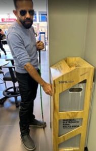 Foto Vertical de Felipe Monteiro, conhecendo o coletor para descarte de produtos eletrônicos na loja Tim de Osasco para o mapeamento de acessibilidade em maio de 2025. Ele é branco, cabelos pretos, curtos, corte no estilo militar, com as laterais mais curtas que o topo, onde as pontas estão descoloridas. Rosto alongado, testa alta, barba e bigode cheios, também curtos e pretos. Usa suéter azul celeste, calça preta, óculos de sol triangular dourado. Os braços estão flexionados. O direito em diagonal descendente, a mão, com os dedos flexionados, toca um móvel em madeira de pinus, encostado na parede de cor areia, em um nicho formado por uma projeção da mesma. O móvel serve de cobertura e escoadouro para um contêiner, uma caixa plástica branca no interior, que recebe descarte de eletrônicos. O braço esquerdo está flexionado paralelo ao dorso, com a mão, na altura do peito, segurando uma bengala verde, que toca o piso entre os pés. As pernas alongadas em paralelo, os pés plantados no piso. O direito, levemente inclinado para fora. A esquerda e por trás de Felipe, parte de uma cadeira e uma mesa. Ao fundo, parte de uma pessoa caminhando pelo pátio e de fachadas de outras lojas. ROTEIRO DE AUDIODESCRIÇÃO: Nadia Moreno CONSULTORIA: Felipe Monteiro 