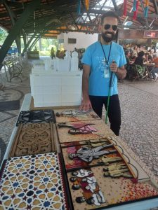 Foto vertical de Felipe Monteiro, ao lado da maquete tátil, no Terceiro Setembro Inclusivo, do Museu da Vida, na FIOCRUZ, em setembro de 2025.
Ele é branco, cabelos pretos, curtos, com corte no estilo militar, com as laterais mais curtas que o topo. Rosto alongado, testa alta, sobrancelhas, barba e bigode também pretos. 
Usa camiseta de manga curta azul, com estampa gráfica em branco, calça preta, tênis preto com solado branco, crachá pendurado no pescoço e óculos de sol triangular dourado. Braço direito alongado, a mão toca em uma das pranchas táteis, com imagens em alto relevo, sobre uma mesa a direita. O braço esquerdo flexionados a frente do corpo, paralelo ao dorso. A mão, do mesmo lado, na altura do peito, segura uma bengala verde. Pernas alongadas. A esquerda em abertura frontal, um pouco à frente da direita. Ele sorri, mostrando dentes brancos e alinhados. 
A esquerda, sobre uma mesa comprida, em branco, a maquete do palácio da FIOCRUZ e diversas pranchas táteis em alto relevo, com detalhes de partes do castelo, dispostas em duas fileiras. Na esquerda, o piso da varanda do terceiro andar, uma parte da parede 
e um dos vitrais. Na direita pessoas representadas no painel que fica no centro de recepção, em frente a arena.
Por trás de Felipe, da esquerda para a direita, o piso em pedras portuguesas brancas e pretas, parte a estrutura de metal do galpão aberto, o pátio da cantina, com diversas pessoas sentadas no entorno de mesas. Ao fundo, parte um terreno arborizado, parte de uma construção de parede branca e a estrutura que delimita o espaço de trabalho da cantina em marrom. Acima parte da estrutura metálica de sustentação do telhado em telhas de barro, com a parte central em policarbonato transparente.  

ROTEIRO DE AUDIODESCRIÇÃO: Nadia Moreno
CONSULTORIA:  Felipe Monteiro
